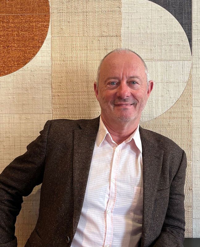 Un homme en blazer marron et chemise rose sourit devant un mur à motifs aux tons neutres.