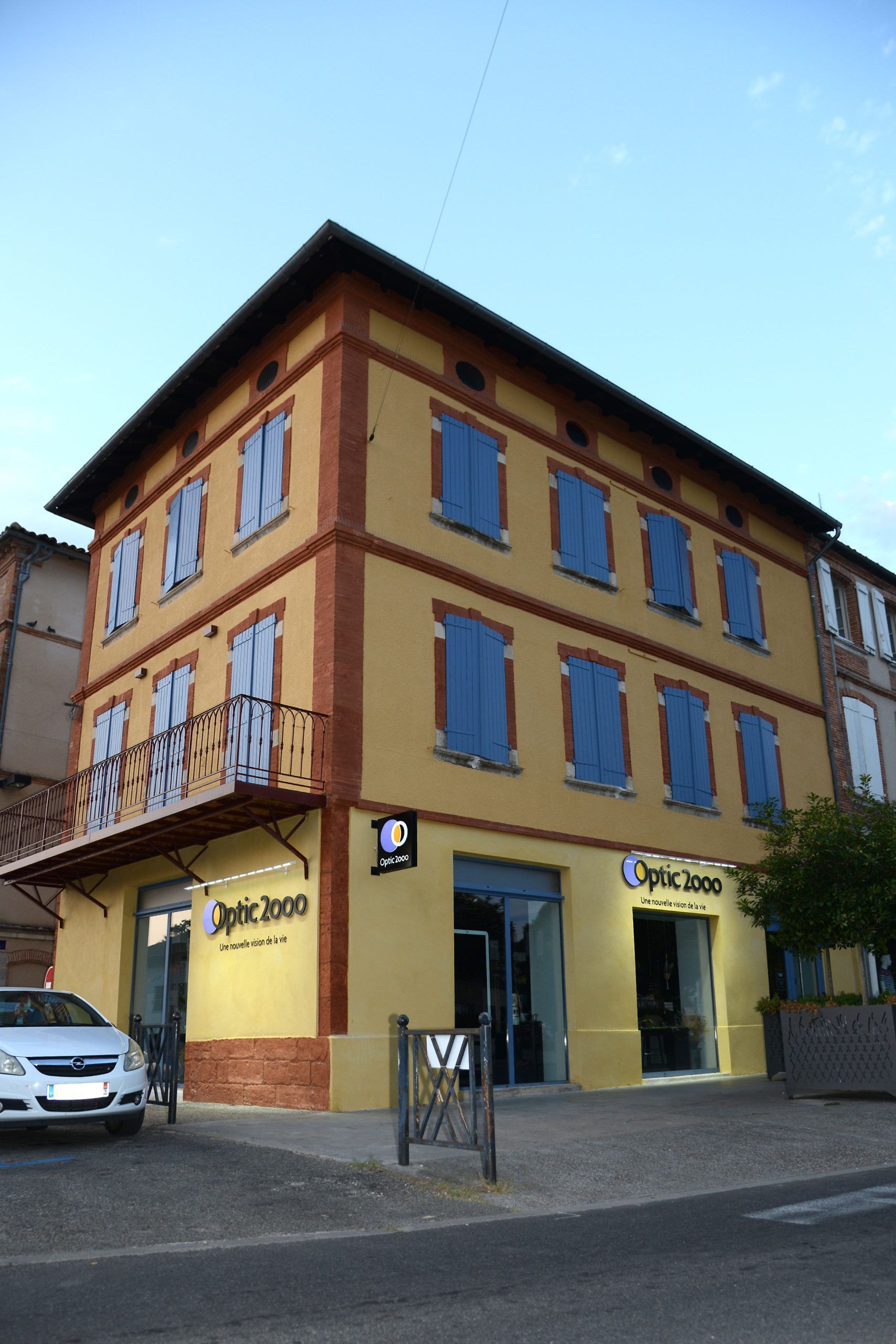 Bâtiment de trois étages avec volets bleus et enseigne Optic 2000 ; façade en briques jaunes.