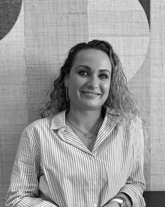 Une femme aux cheveux bouclés sourit, vêtue d'une chemise à rayures et de bijoux, posant devant un mur texturé.