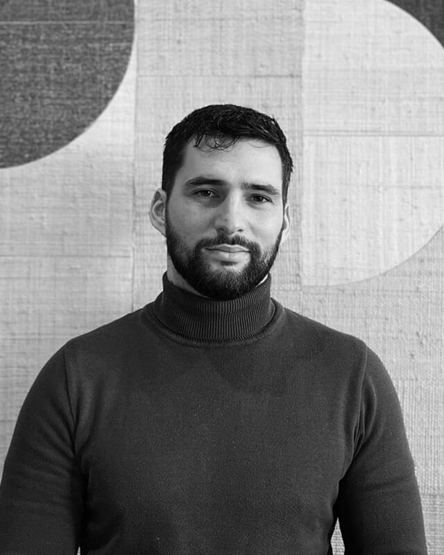 Un homme barbu, vêtu d'un pull à col roulé foncé, se tient devant un mur à motifs.