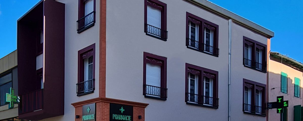 Un immeuble avec une enseigne de pharmacie à l'angle. Des boiseries brunes et un grand auvent. Journée ensoleillée.