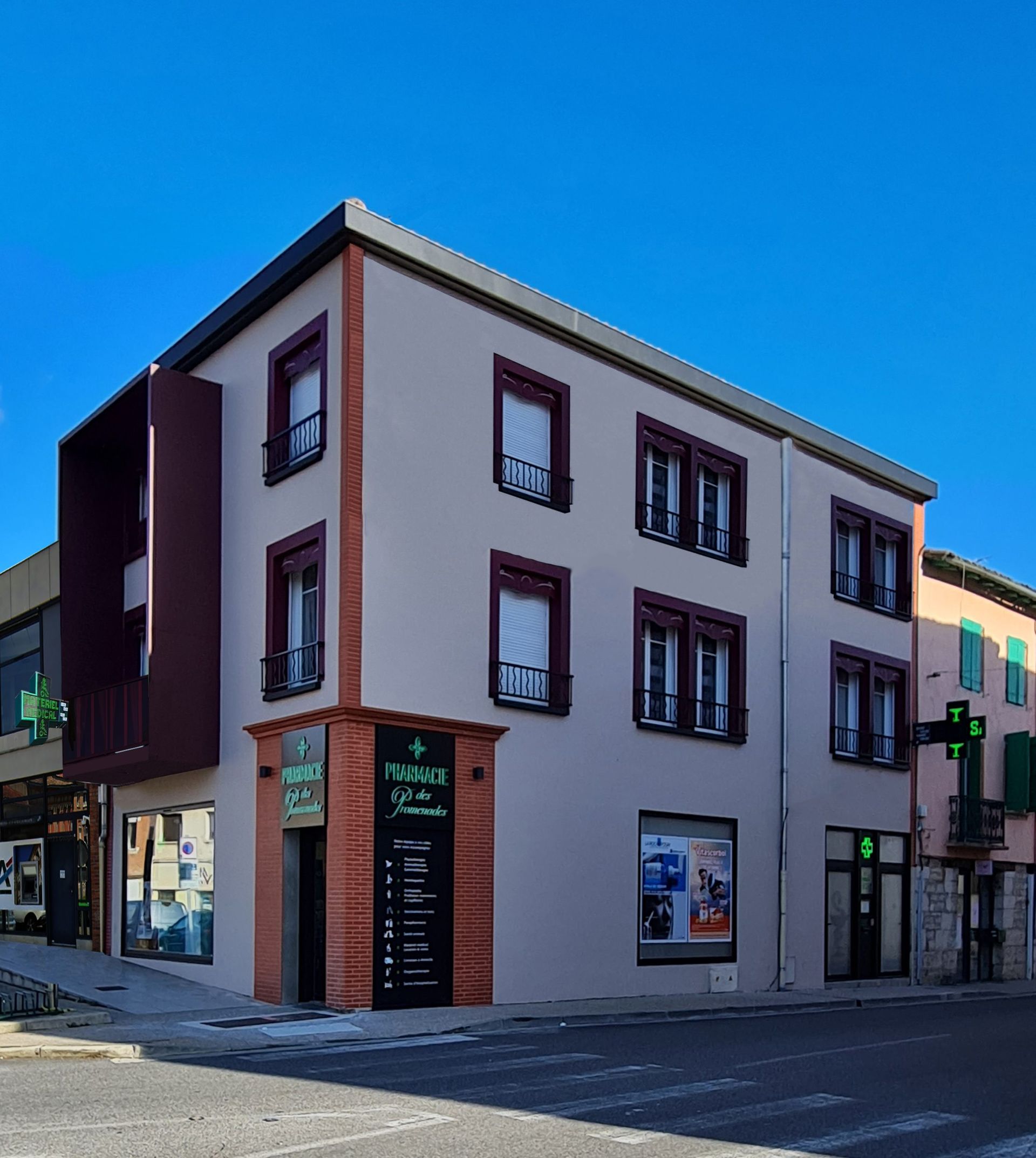 Un bâtiment de trois étages abritant une pharmacie, avec des touches de rouge foncé et une enseigne sur une rue sous un ciel bleu.