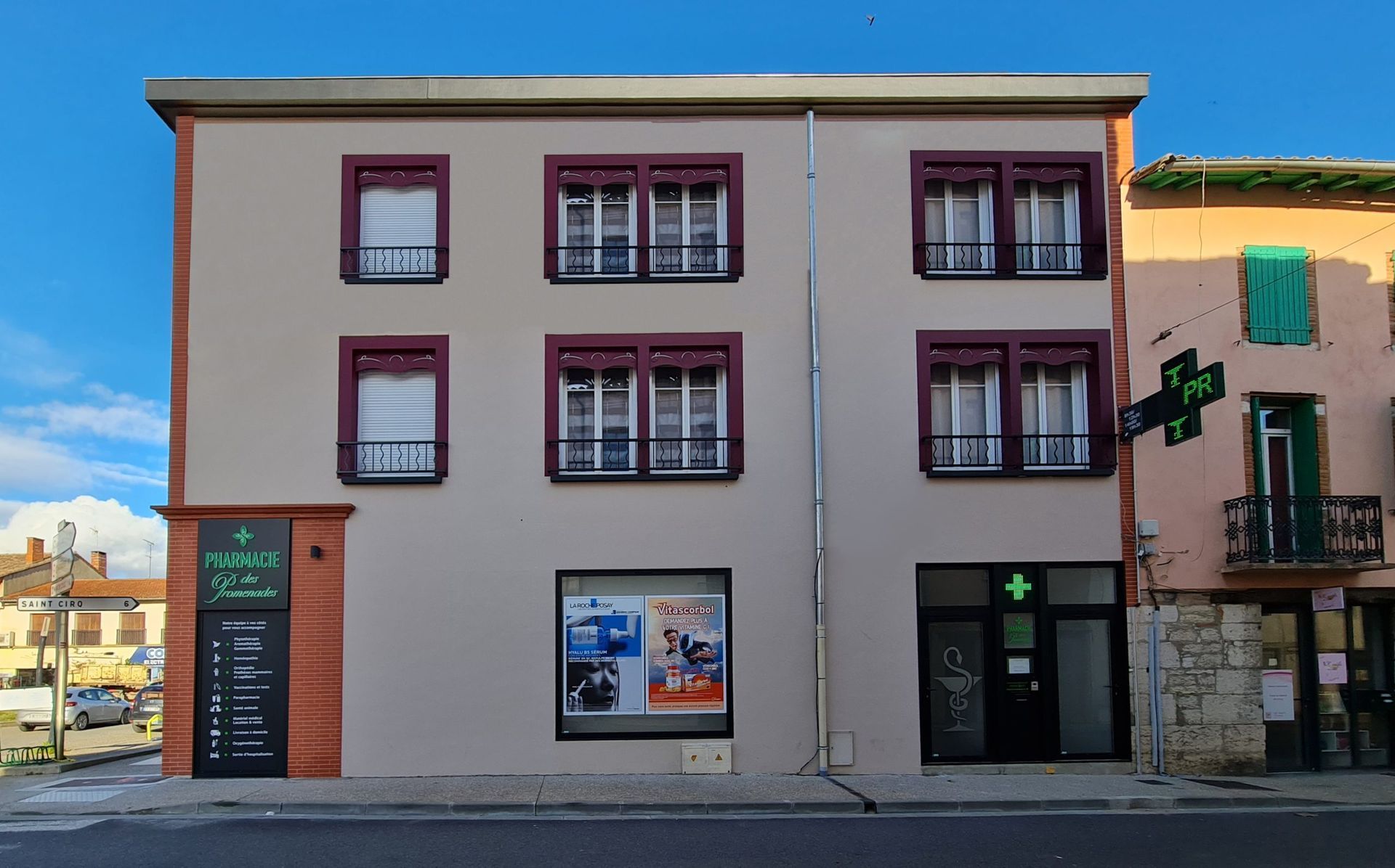 Pharmacie de deux étages avec une croix verte et une enseigne. Les fenêtres ont des encadrements bordeaux.
