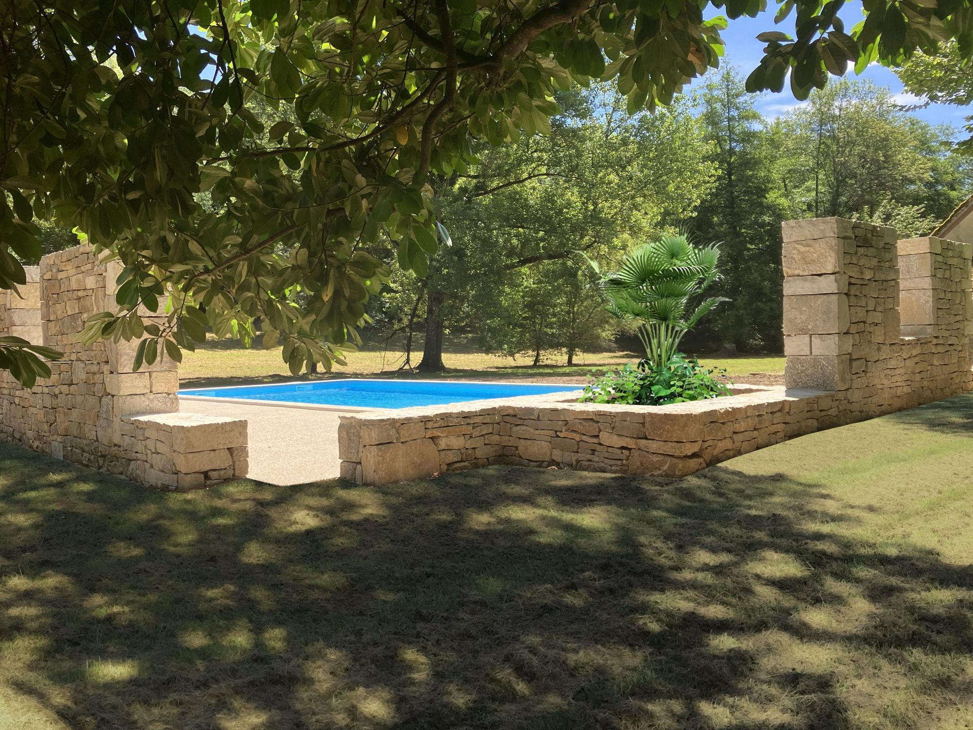 Piscine entourée de murs en pierre, dans un jardin gazonné, sous la canopée d'un arbre. Eau d'un bleu éclatant, plantes vertes et arrière-plan ensoleillé.