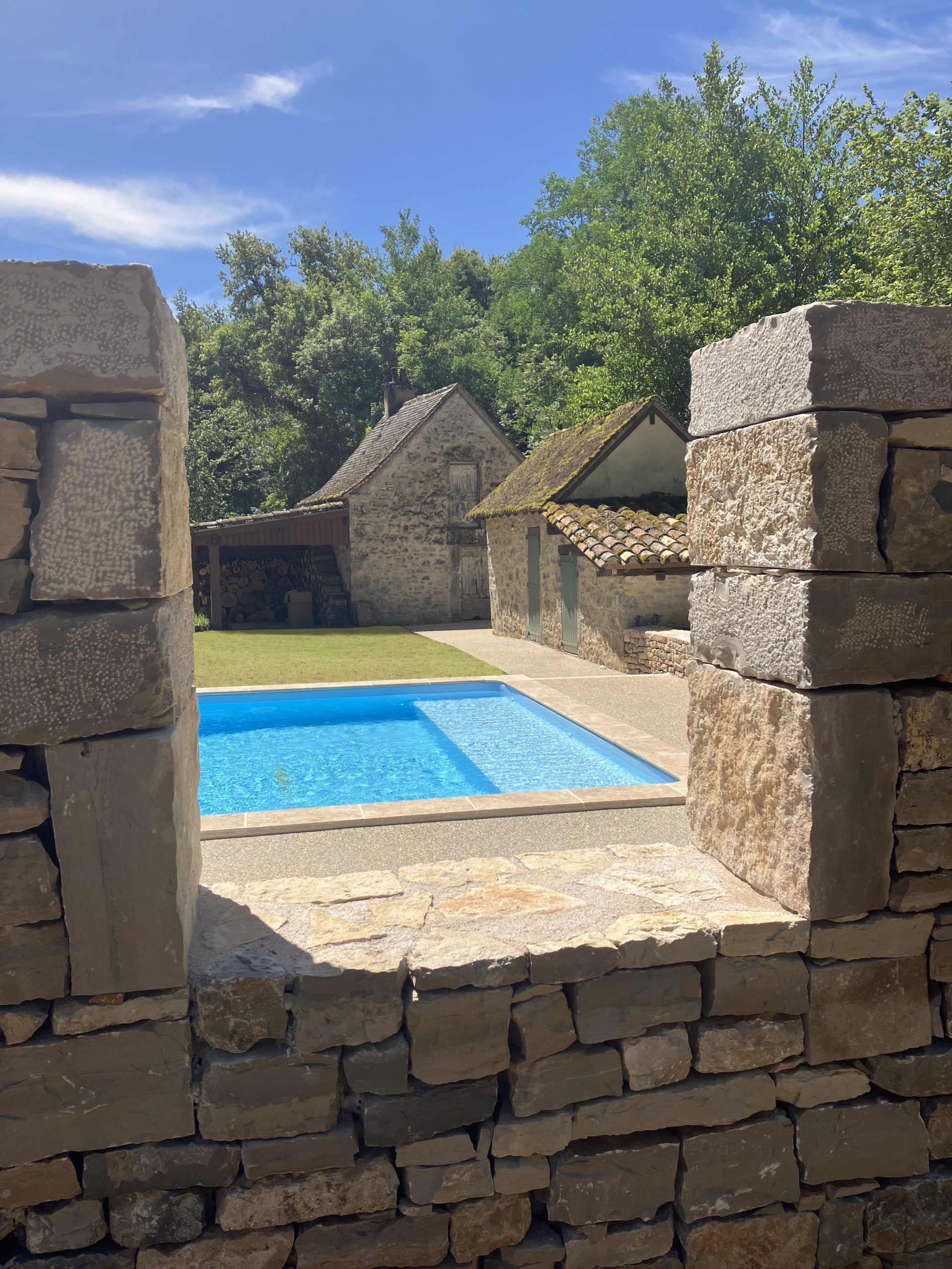 Un mur de pierre encadrant une piscine bleue, des bâtiments en pierre et des arbres verts sous un ciel bleu.