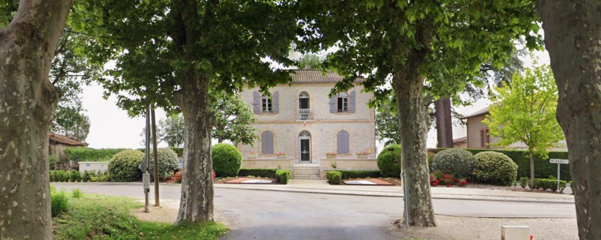 Un bâtiment en pierre encadré par des arbres, dans une rue bordée d'arbres.