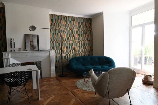 Salon avec canapé turquoise, mur à motifs, cheminée et grande fenêtre.