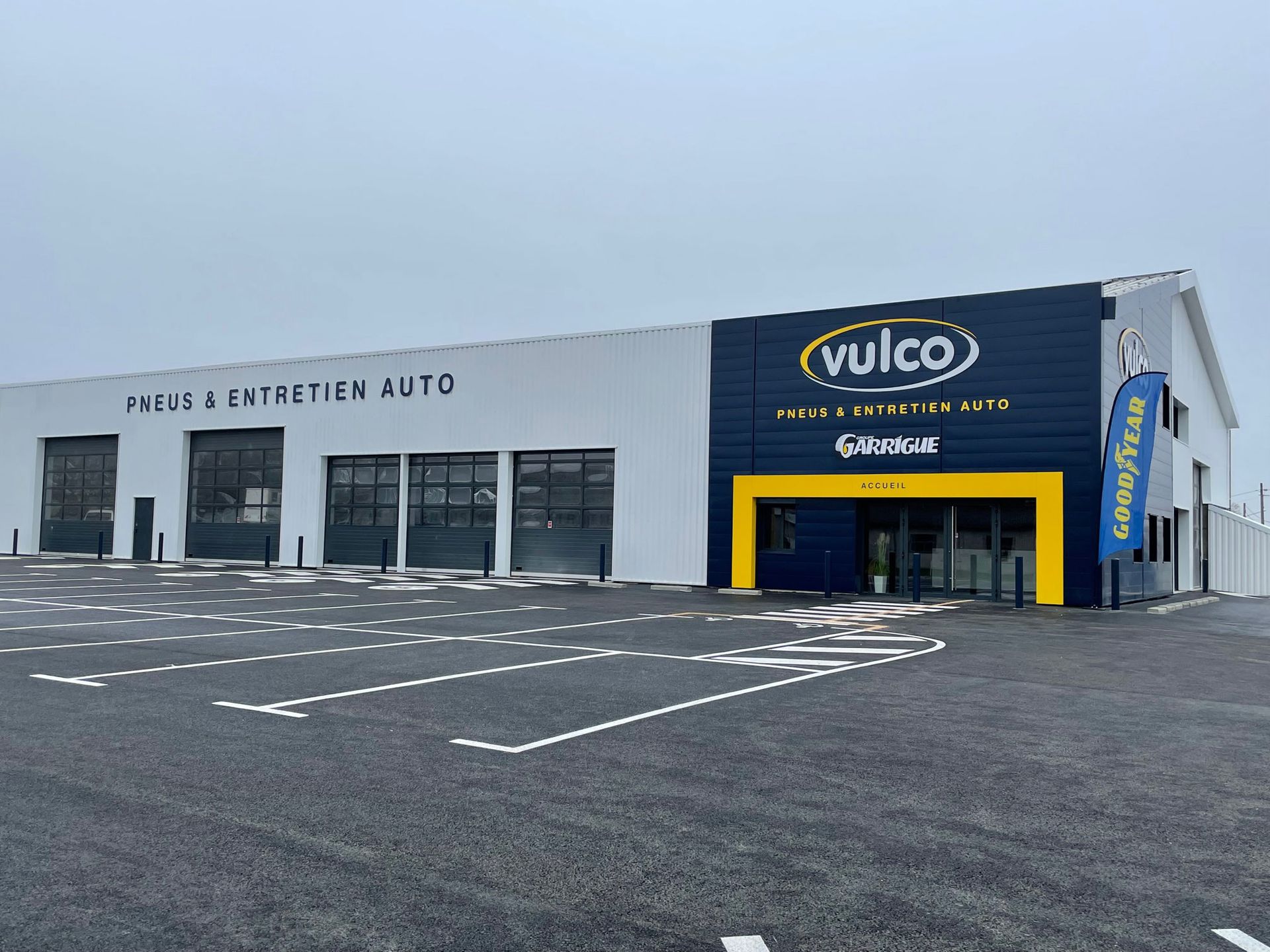 Extérieur d'un garage automobile Vulco. Bâtiment blanc et bleu marine avec portes de garage, touches de jaune et enseigne.