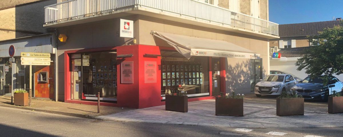 Un bâtiment aux accents rouges, doté de grandes fenêtres et d'un auvent rétractable. Plusieurs plantes en pot bordent le trottoir.
