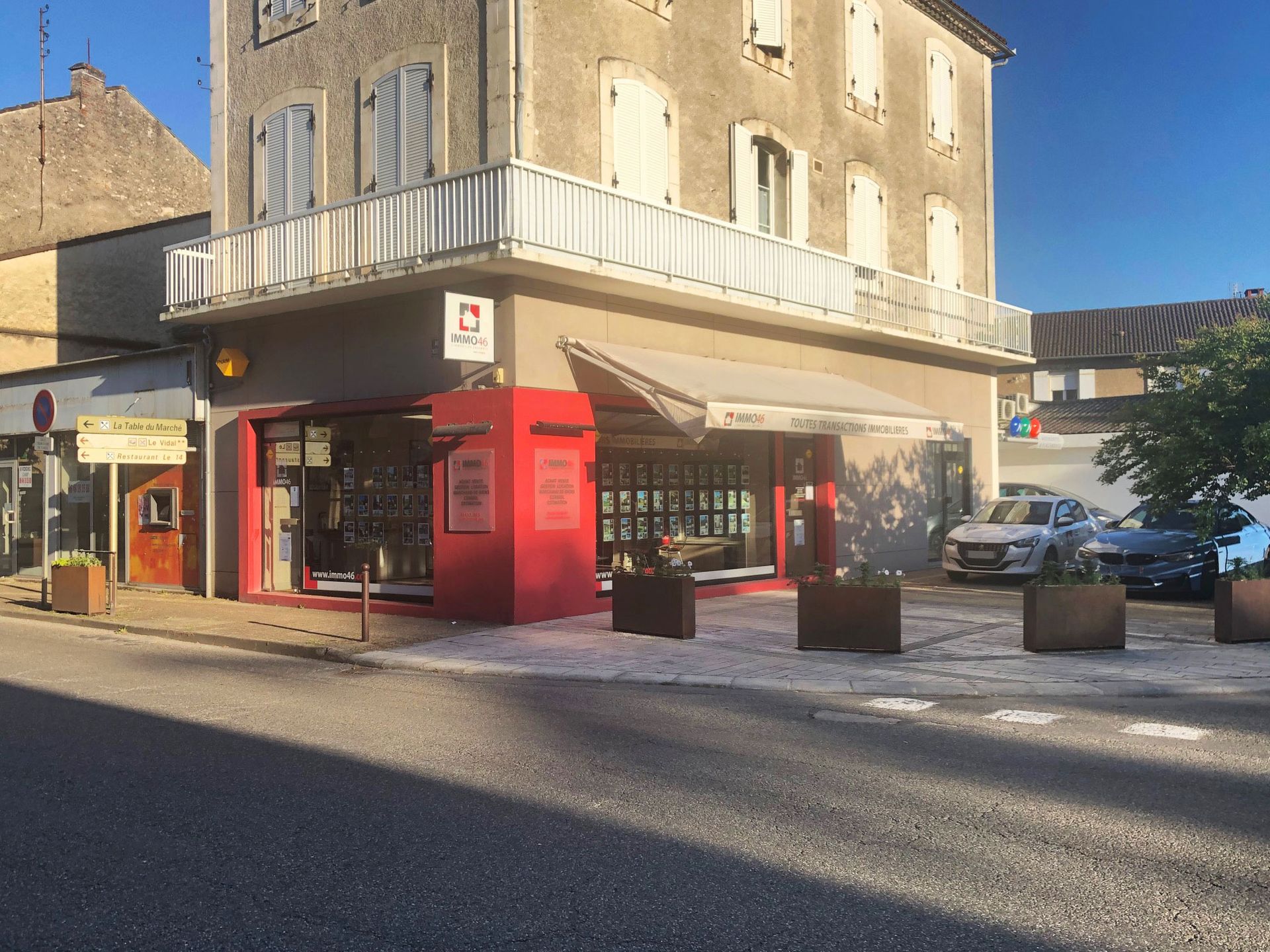 Vue de rue d'un immeuble de deux étages avec des commerces au rez-de-chaussée. Une boutique rouge est dotée d'un auvent. Des voitures sont stationnées à proximité.