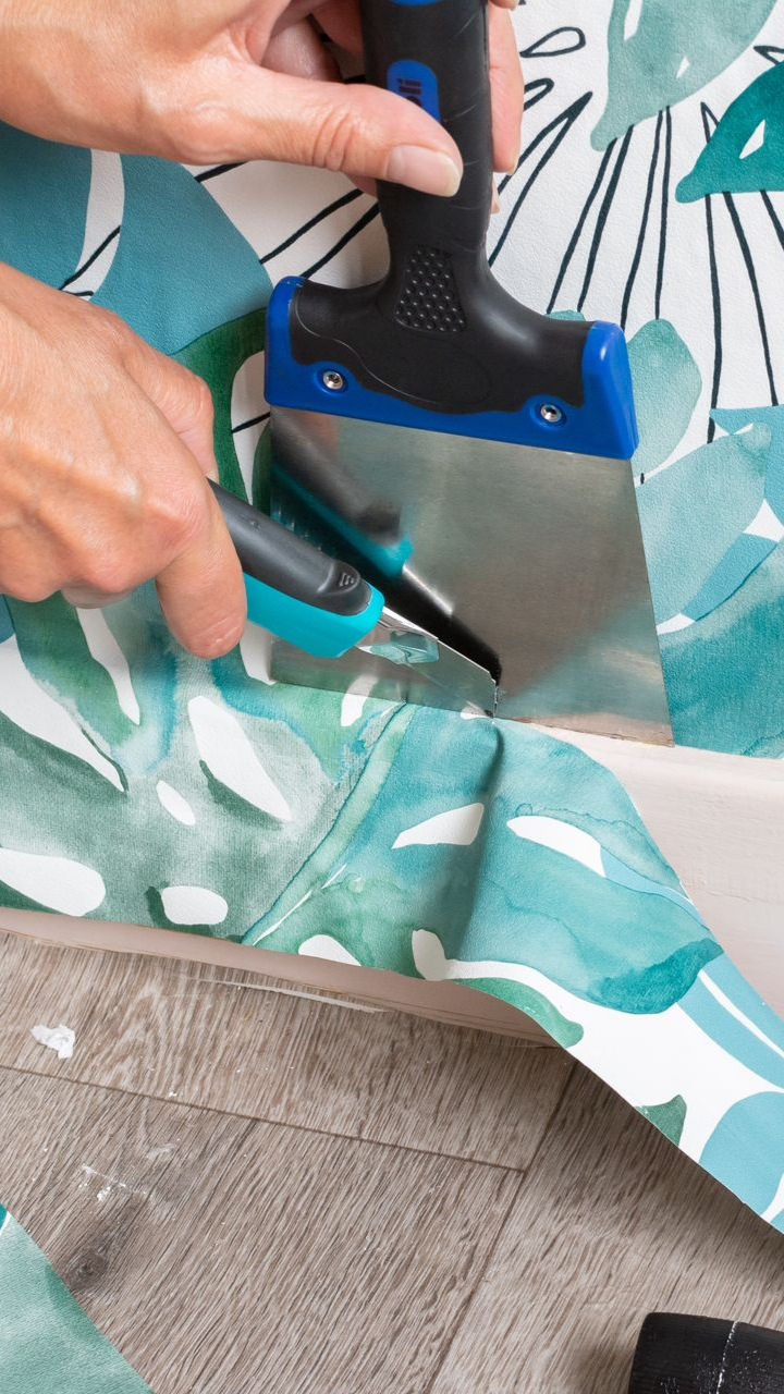 Personne utilisant un grattoir et un couteau utilitaire pour couper du papier peint près d'une surface, motif floral vert et bleu.