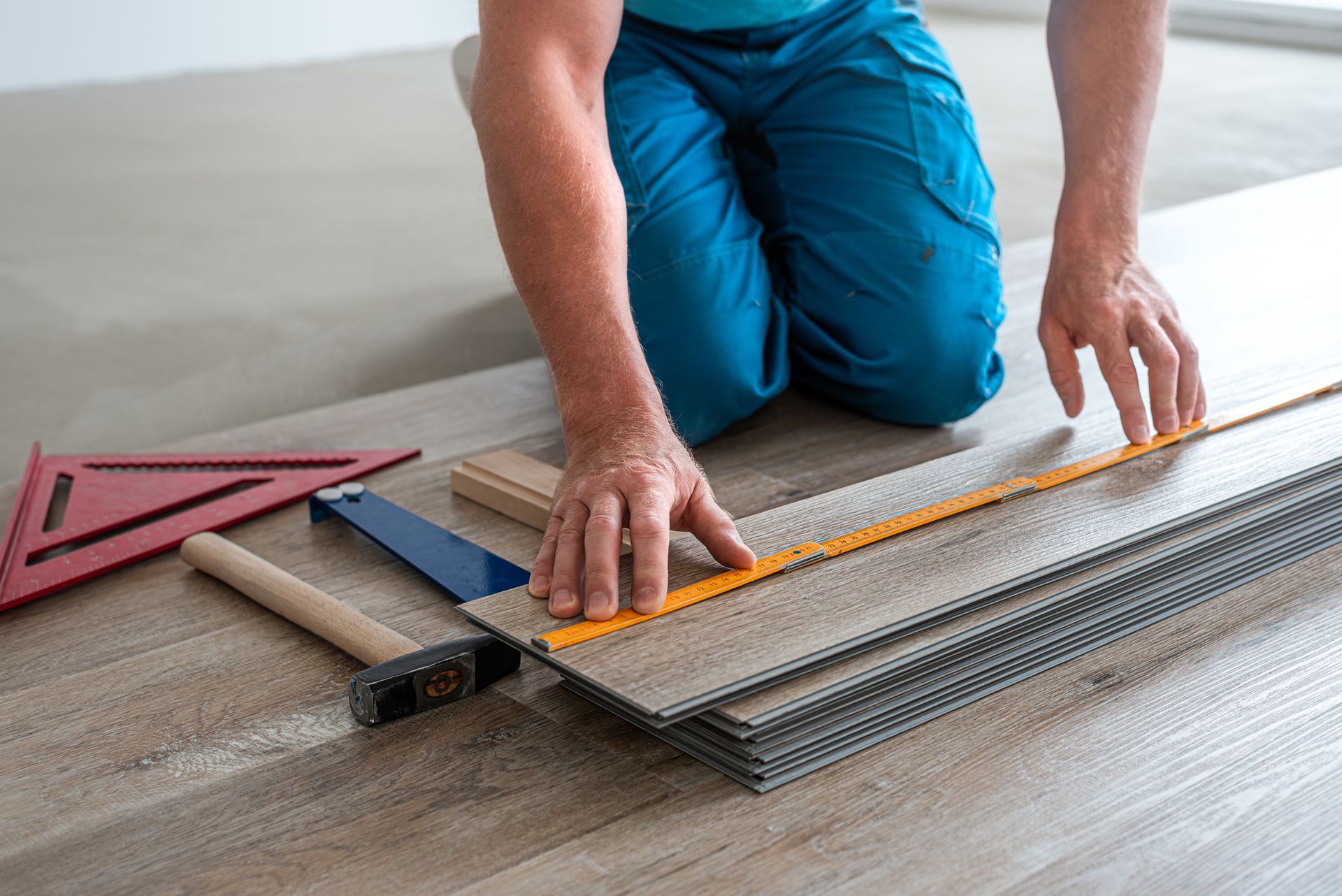 Personne installant un revêtement de sol, mesurant avec une règle, outils à proximité : marteau, équerre et blocs.