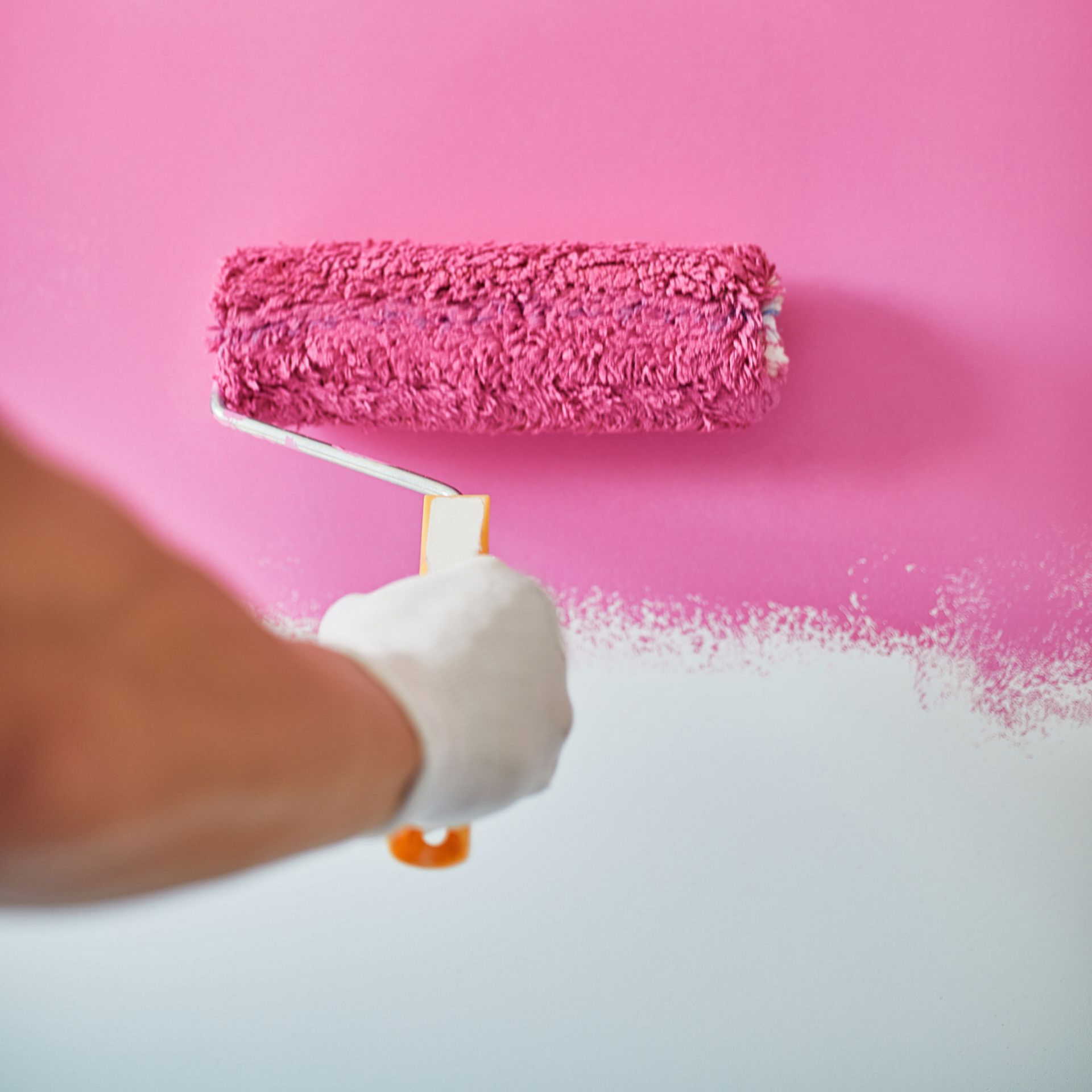 Main portant un gant blanc utilisant un rouleau à peinture rose pour peindre un mur en rose.