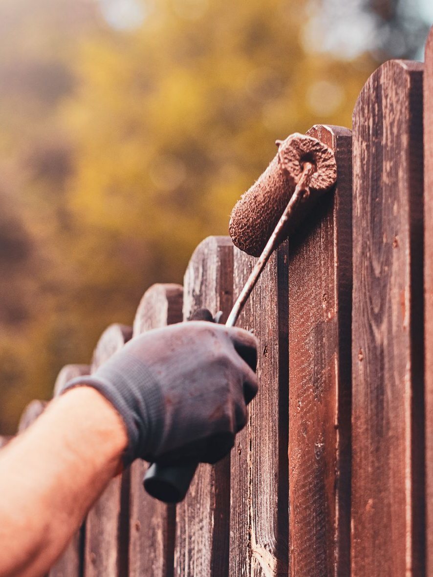 Main dans un gant noir utilisant un rouleau à peinture pour teindre une clôture en bois, à l'extérieur.