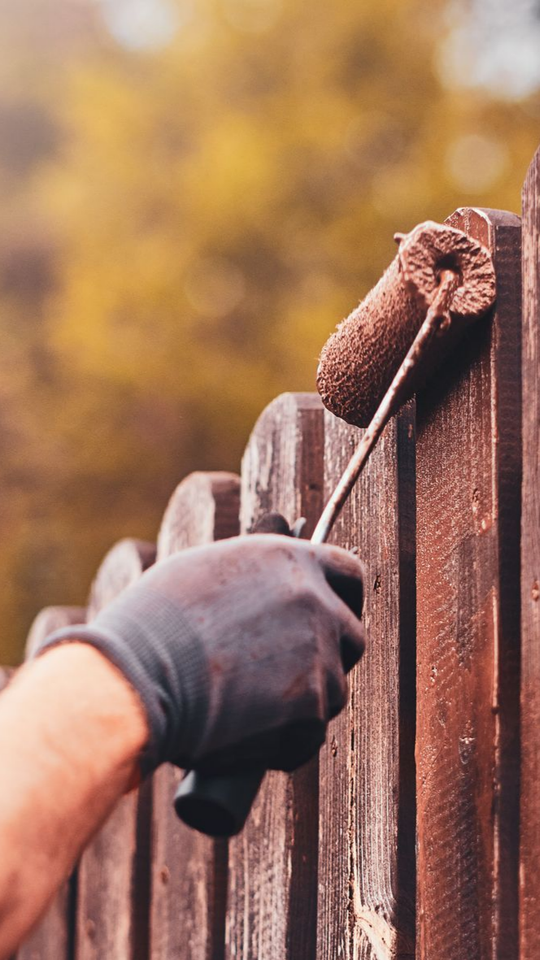 Une personne portant un gant peint une clôture en bois avec un rouleau à peinture, à l'extérieur.