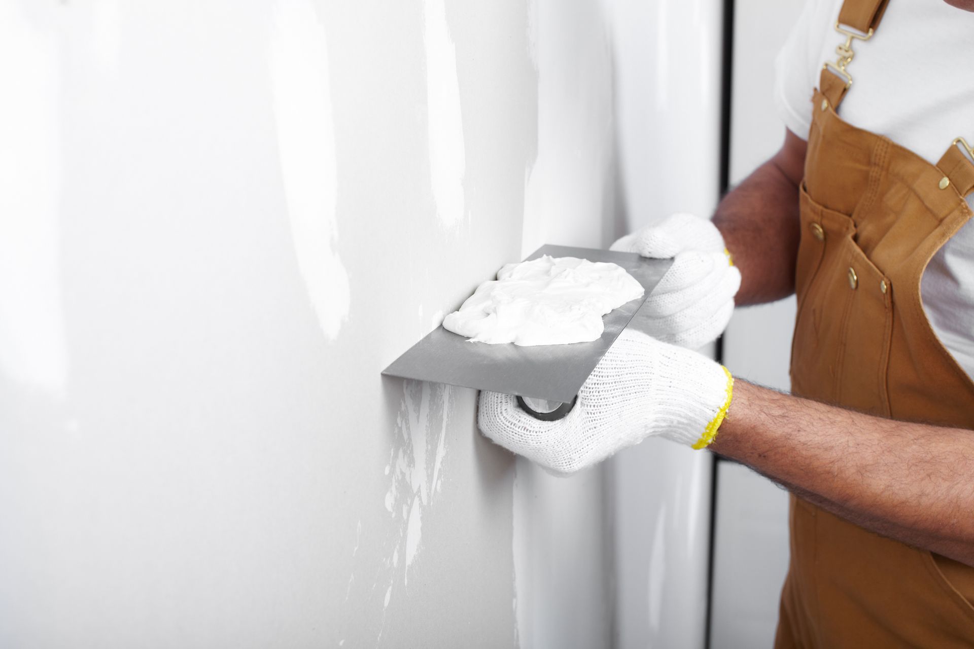 Une personne portant des gants, appliquant du mastic sur un mur blanc avec un couteau à mastic en métal.