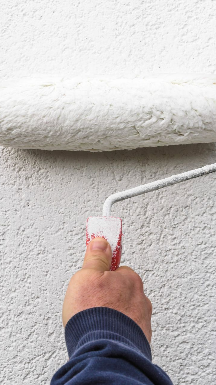 Une main tenant un rouleau à peinture appliquant de la peinture blanche sur un mur texturé.