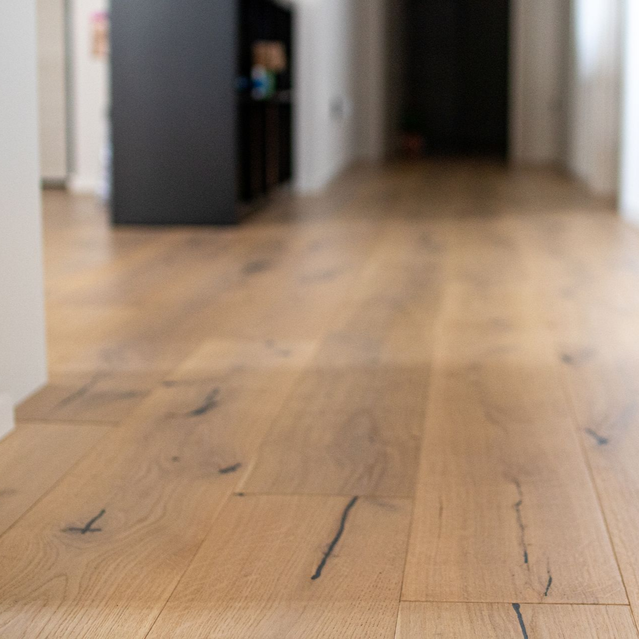 Parquet en bois clair dans un couloir, avec une armoire noire et une porte floue en arrière-plan.