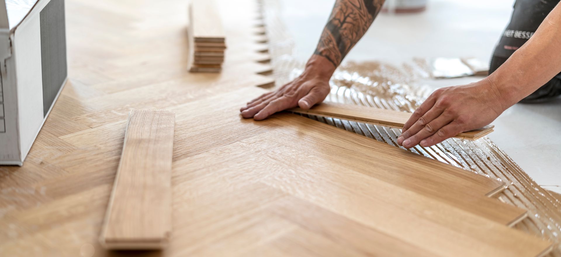 Une personne installant un parquet en bois, appliquant de la colle sur une rangée de planches avec un bras tatoué visible.