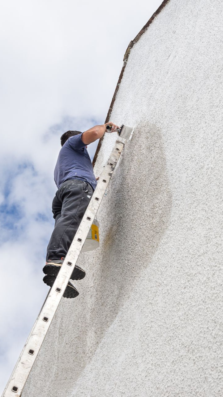 Personne sur une échelle travaillant sur un mur extérieur. Chemise bleue, pantalon gris, ciel nuageux.