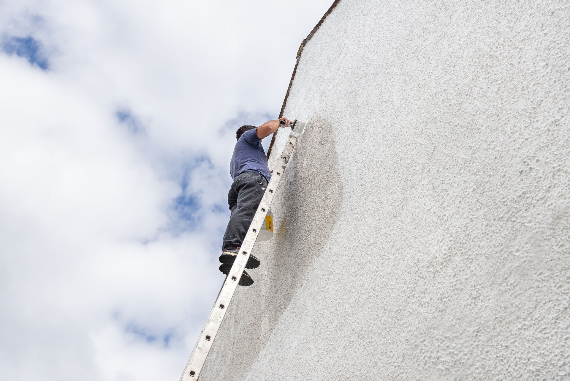 Une personne sur une échelle répare le mur extérieur d'un bâtiment. Ciel et nuages en arrière-plan.