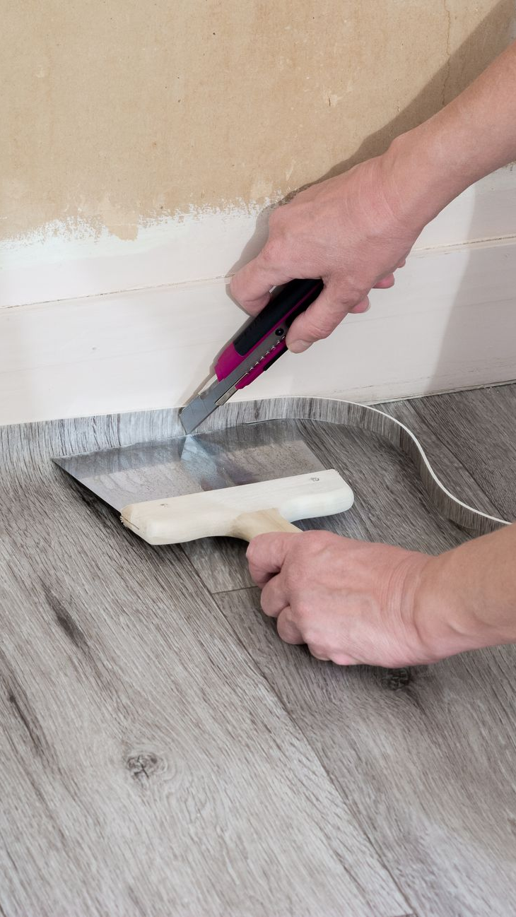 Personne coupant le revêtement de sol avec un couteau utilitaire et le lissant avec un couteau à mastic près d'un mur.