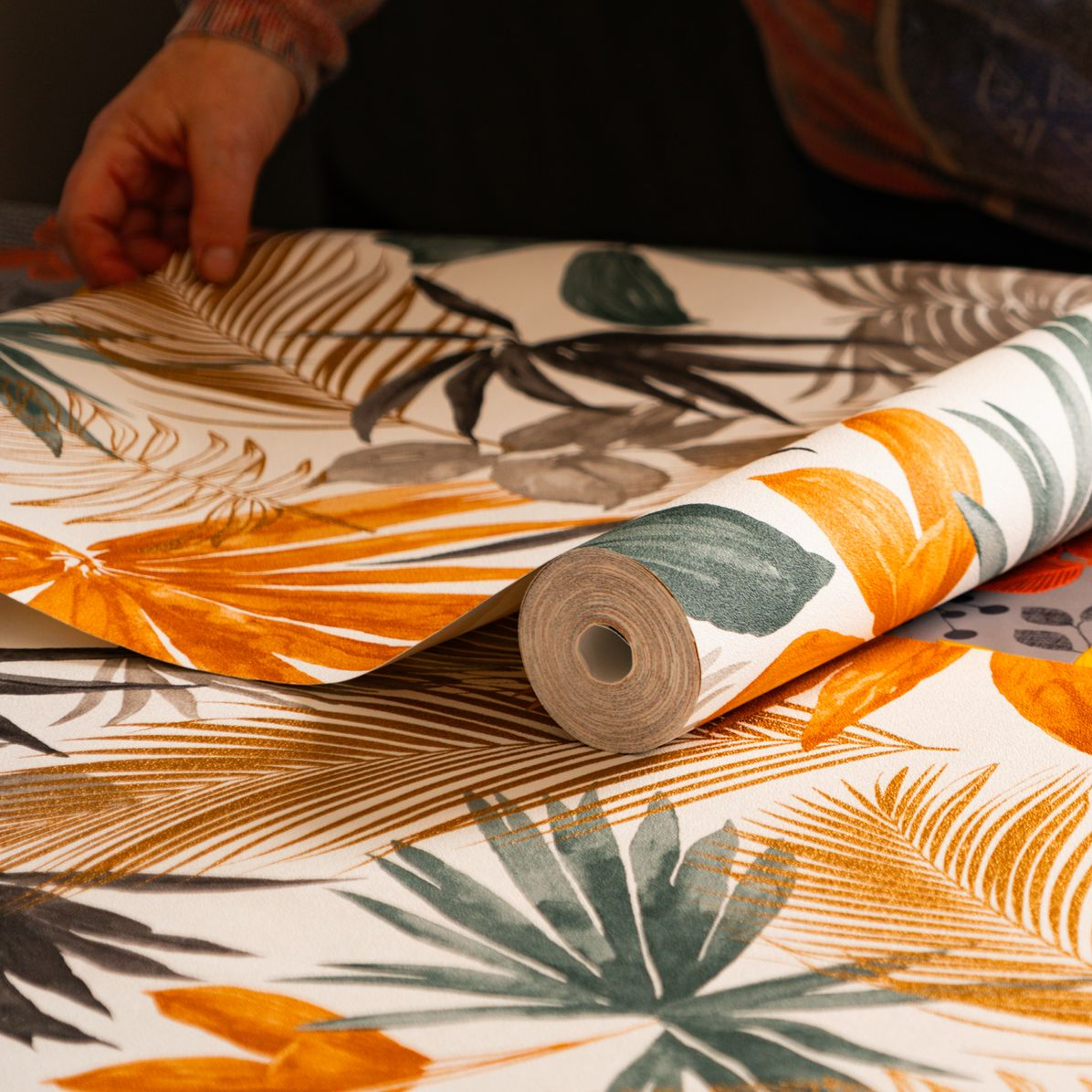 Personne déroulant du papier peint à motif de feuilles tropicales orange, grises et vertes.