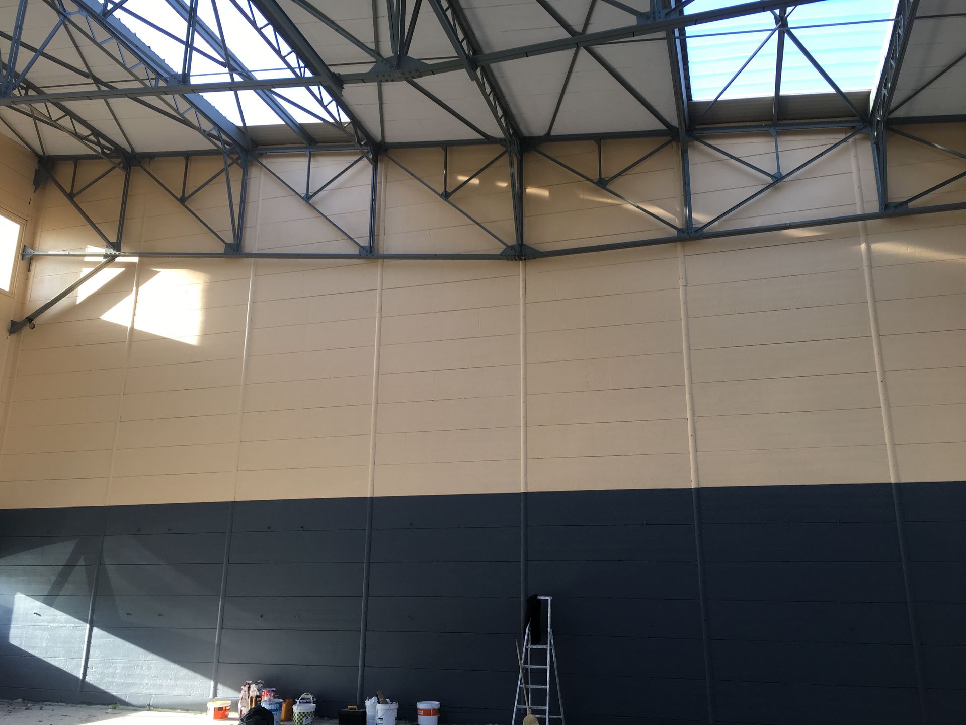 Intérieur d'un grand bâtiment aux poutres métalliques, murs beiges et gris foncé, et une verrière. Une échelle est présente.