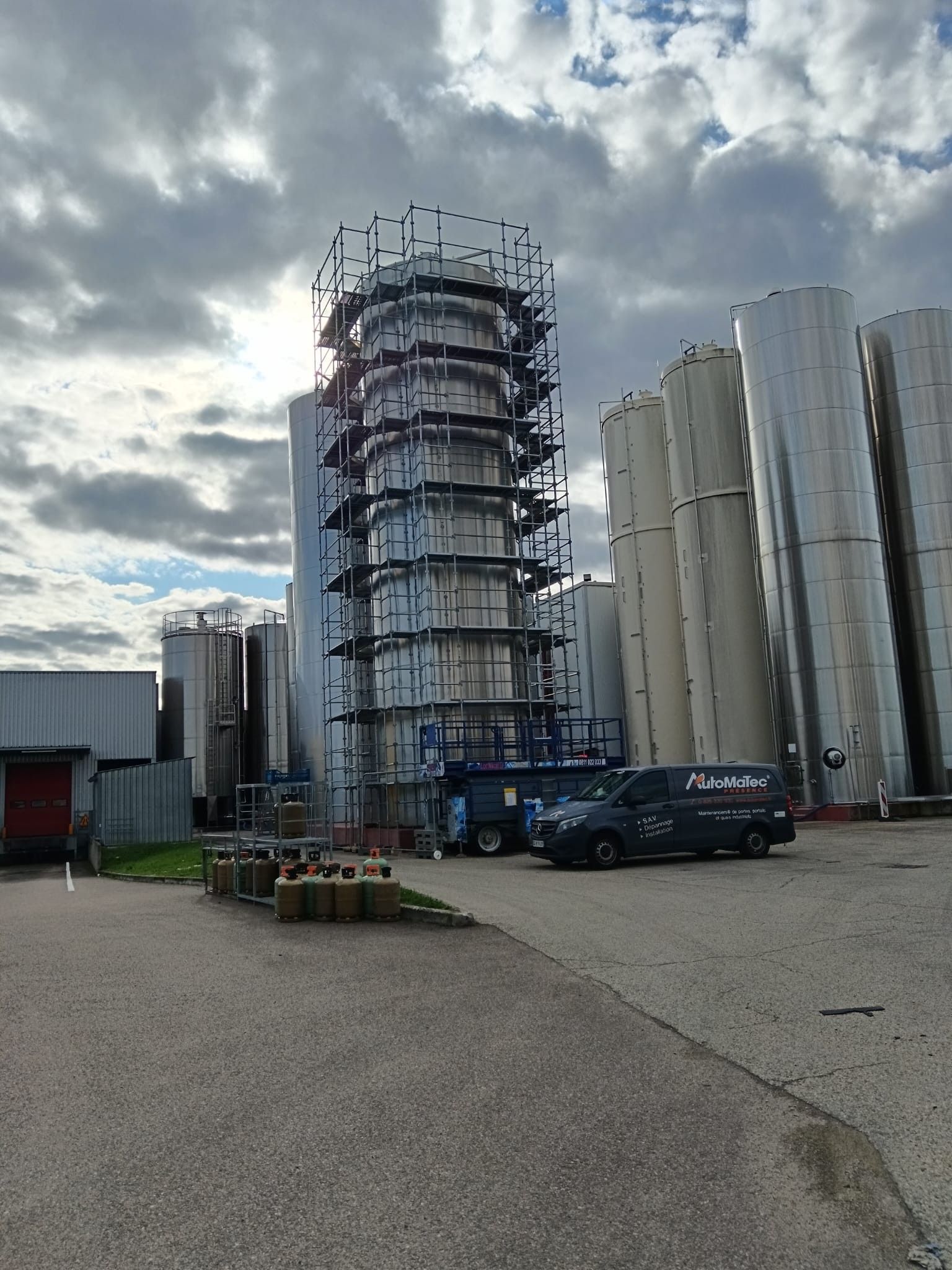 Tour industrielle en construction, entourée d'échafaudages, de réservoirs de stockage et d'un ciel nuageux.