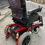 Una silla de ruedas eléctrica roja y negra está estacionada al costado de la carretera.