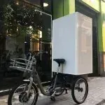 Un triciclo con una caja blanca en la parte trasera está estacionado frente a un edificio.