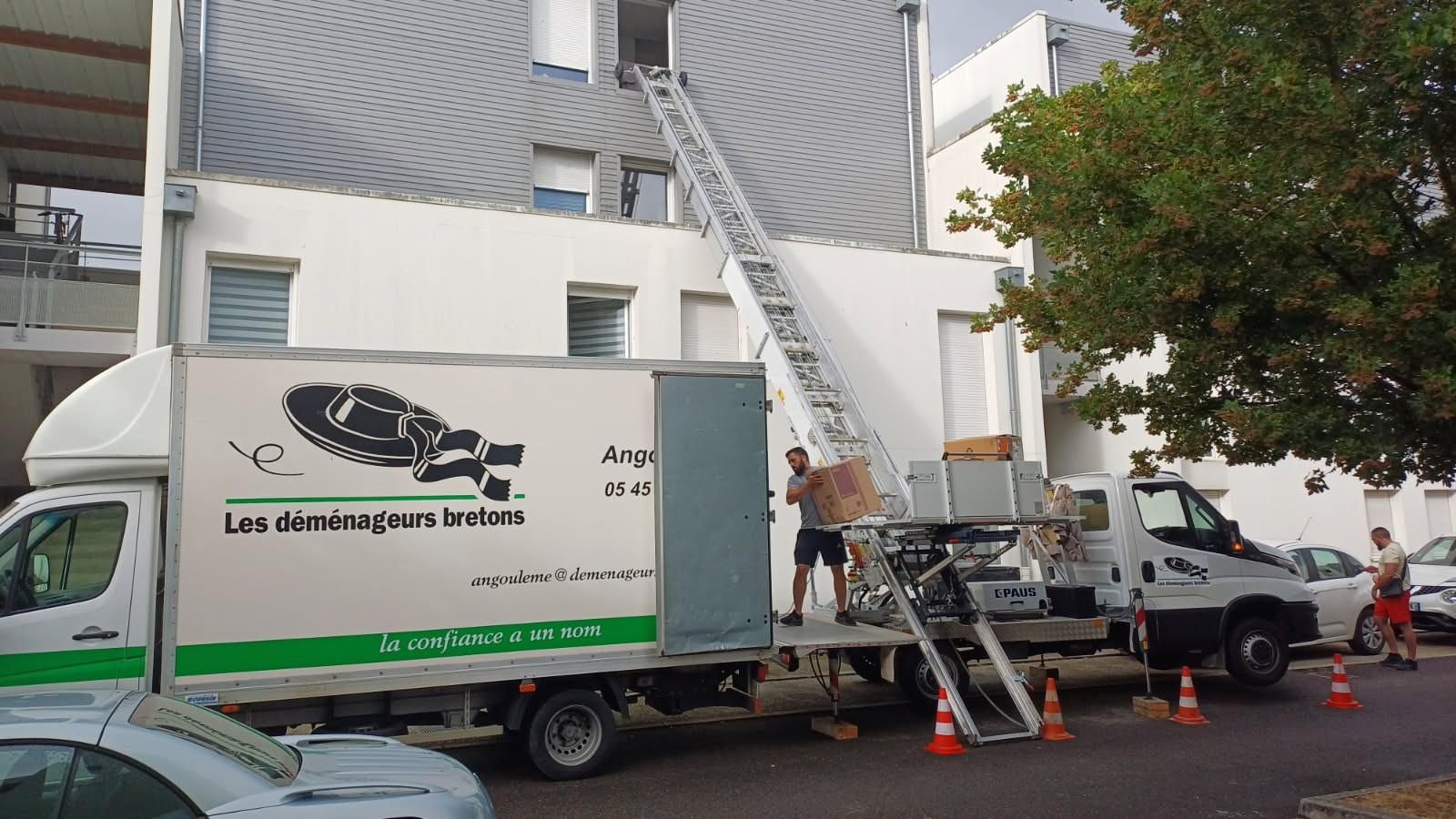 Des déménageurs déchargent des meubles d'un camion à l'aide d'une échelle qui atteint la fenêtre d'un appartement ; des cônes de signalisation sont disposés dans la rue.
