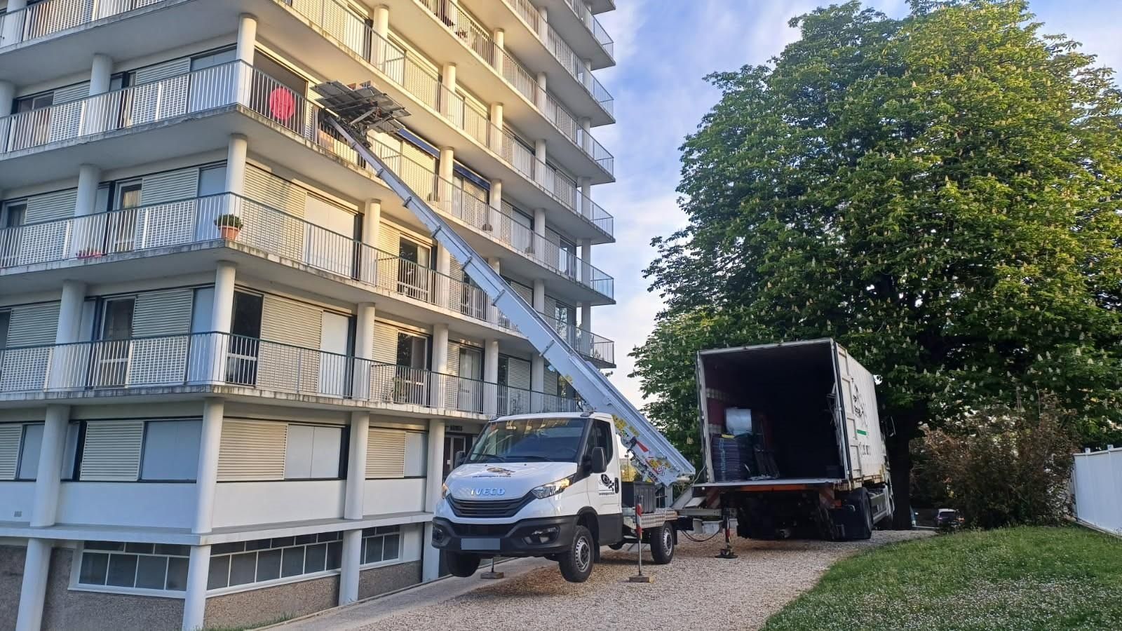 Un camion blanc et une remorque ouverte sont garés à côté d'un immeuble d'appartements, une échelle étant déployée le long de la façade.