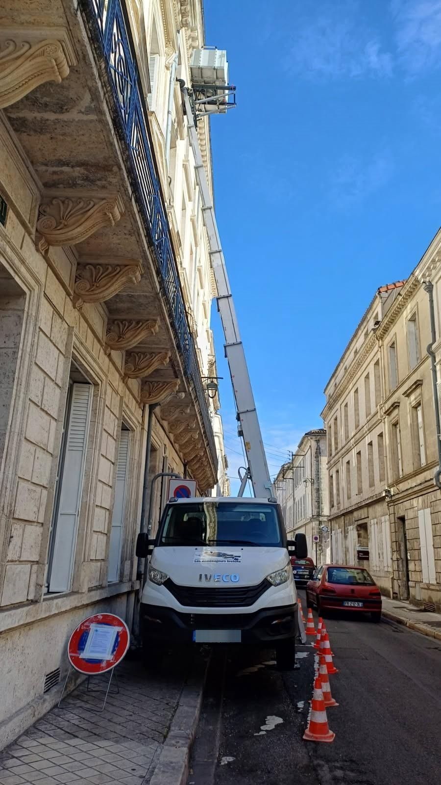 Fourgonnette de service blanche avec échelle déployée, stationnée dans une rue étroite entre de hauts immeubles en pierre, cônes de signalisation à proximité