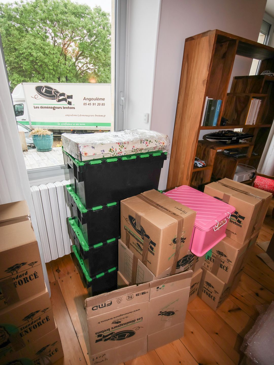 Une personne transporte des cartons empilés dans une pièce lumineuse, avec du matériel de déménagement en arrière-plan.