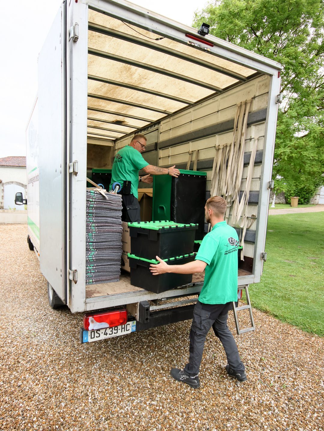 Deux déménageurs chargent des cartons dans un camion de livraison devant une maison d'une rue ensoleillée.