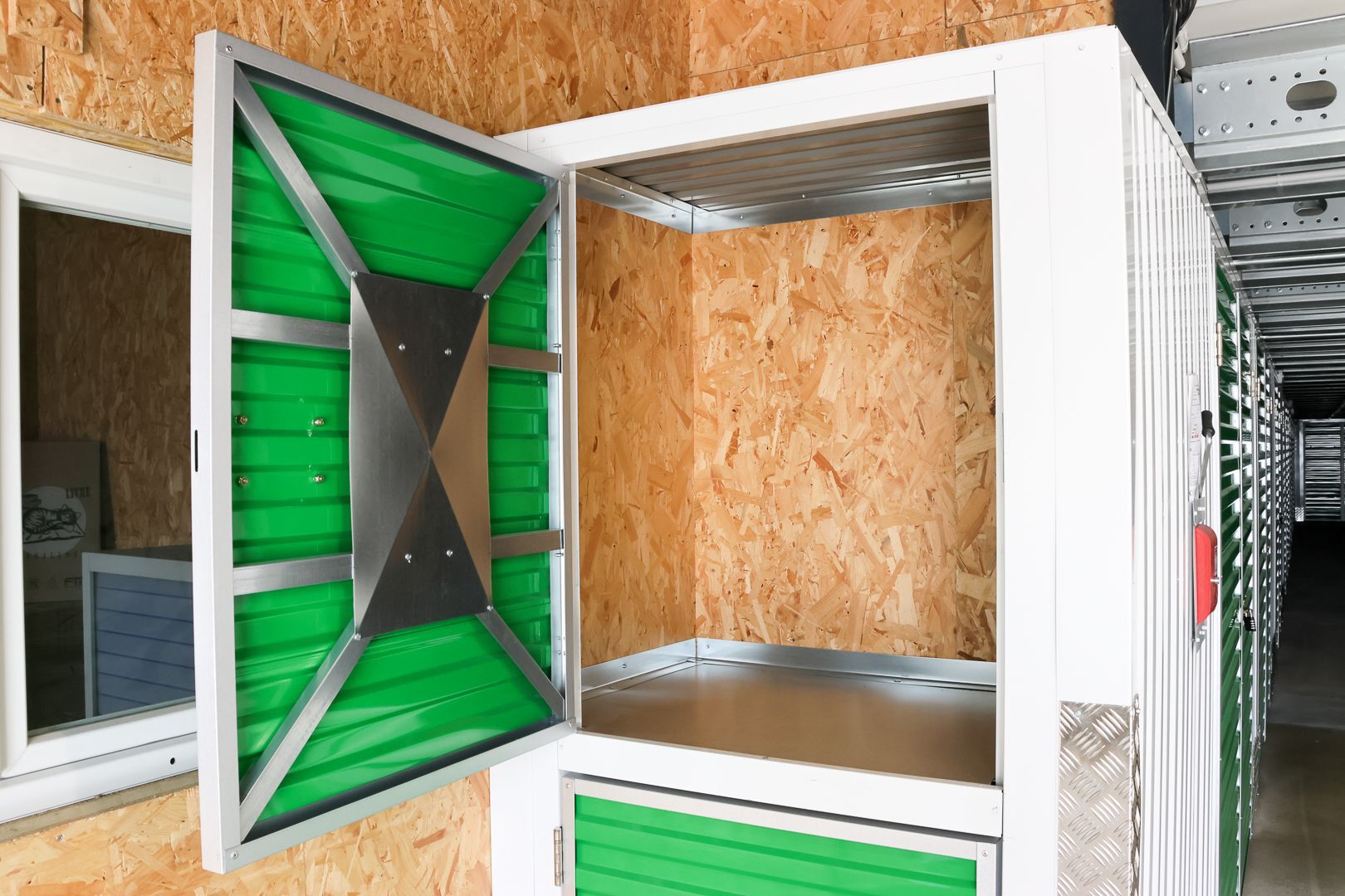 Couloir d'un box de stockage avec portes rouges et objets entreposés près d'un box ouvert, notamment des chaises et un vélo.