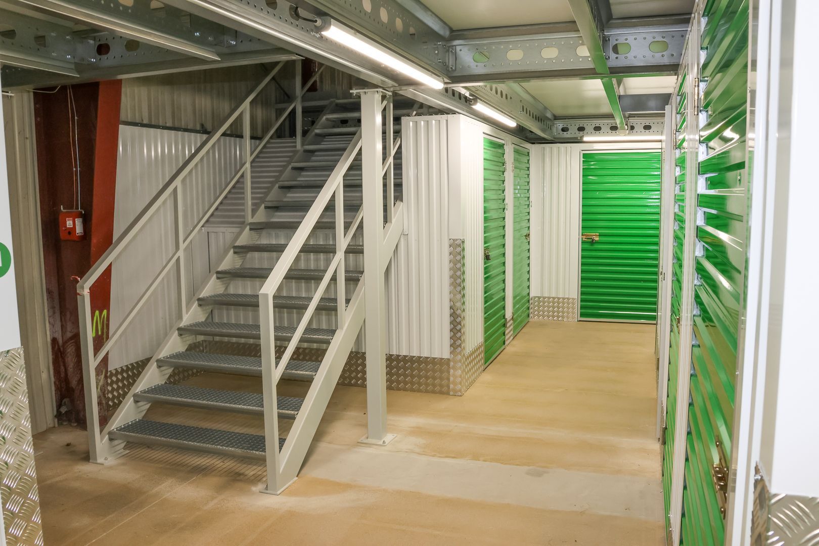 Dans un couloir, une rangée de portes rouges de box de stockage, avec des meubles empilés et un fauteuil roulant près d'un box ouvert.