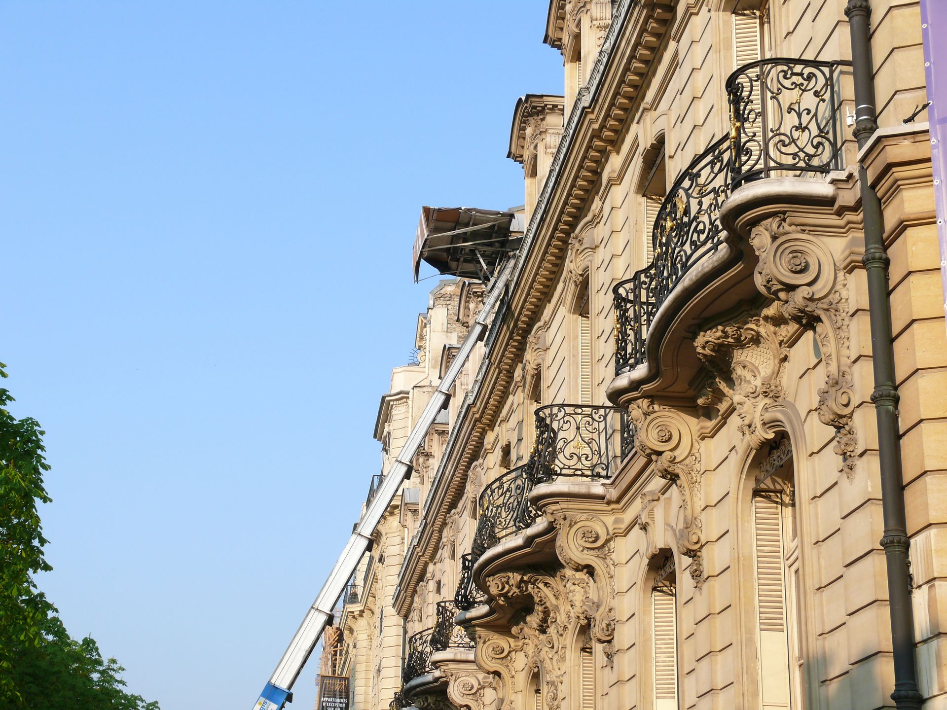 Façade d'un haut bâtiment en pierre avec des balcons ornés se détachant sur un ciel bleu, vue d'en bas