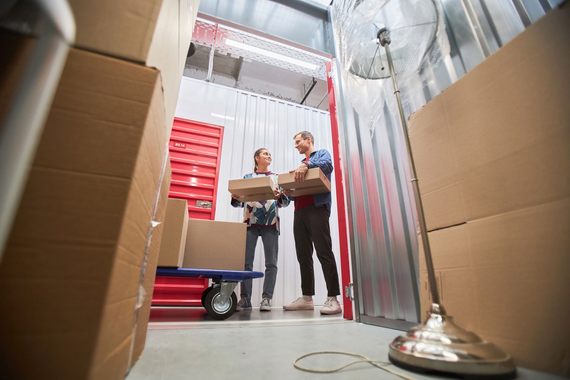 Deux déménageurs transportent des cartons dans une allée d'un entrepôt, avec des bacs rouges et un diable.