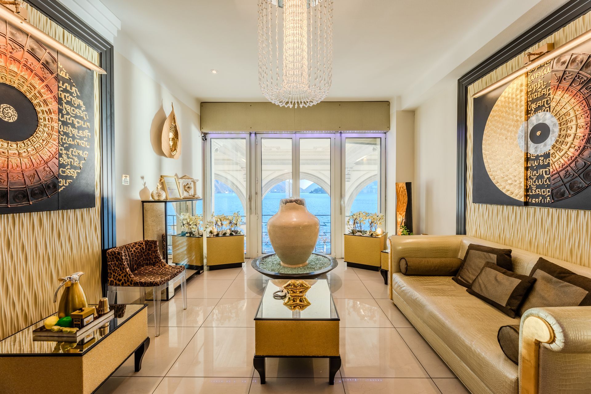 Elegante soggiorno con dettagli dorati, opere d'arte, divano beige, tavolo in vetro e un grande lampadario.