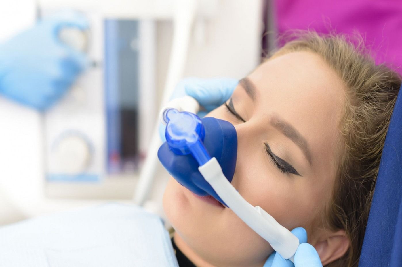 Donna sottoposta a sedazione dentale con una maschera blu per l'ossigeno. Un operatore con guanti blu regola la macchina.