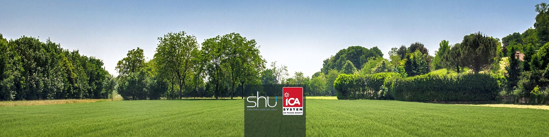 Ampio campo verde con alberi sullo sfondo sotto un cielo azzurro. Un cartello è posizionato al centro del campo.