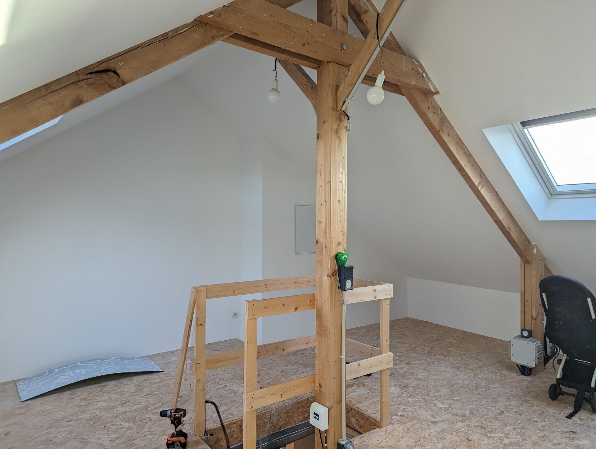 Intérieur des combles avec poutres apparentes, murs blancs, copeaux de bois au sol et une petite lucarne.