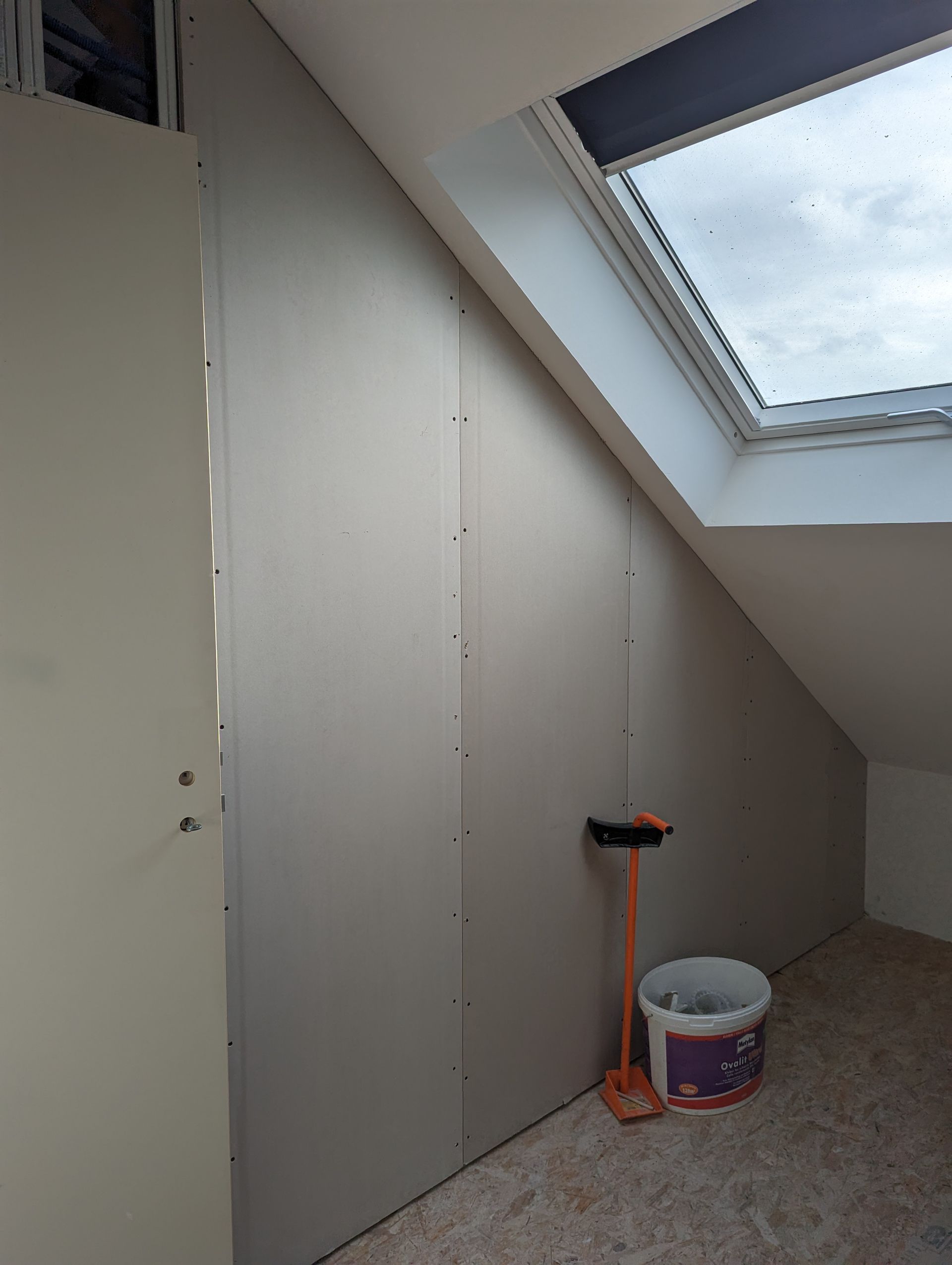 Cloison sèche installée sur un mur incliné dans une pièce mansardée, avec une fenêtre, un balai et un seau.