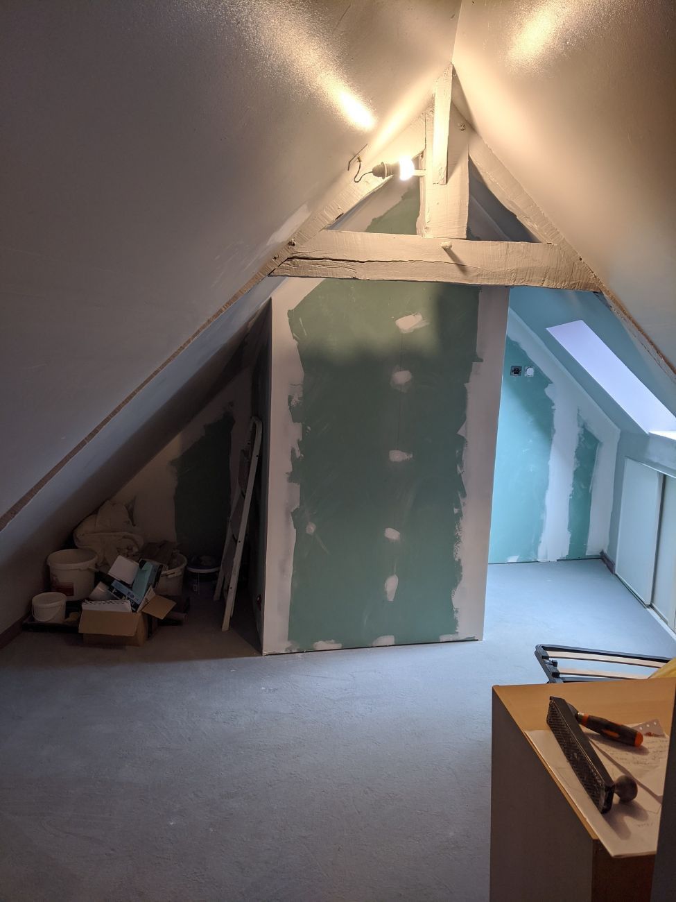 Pièce mansardée en cours d'aménagement avec poutres apparentes, cloisons sèches et matériaux épars.