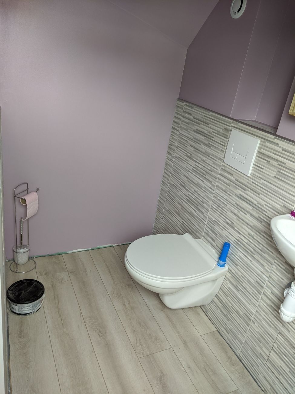 Petite salle de bains avec toilettes et lavabo ; sol clair imitation bois, murs carrelés gris et violets.