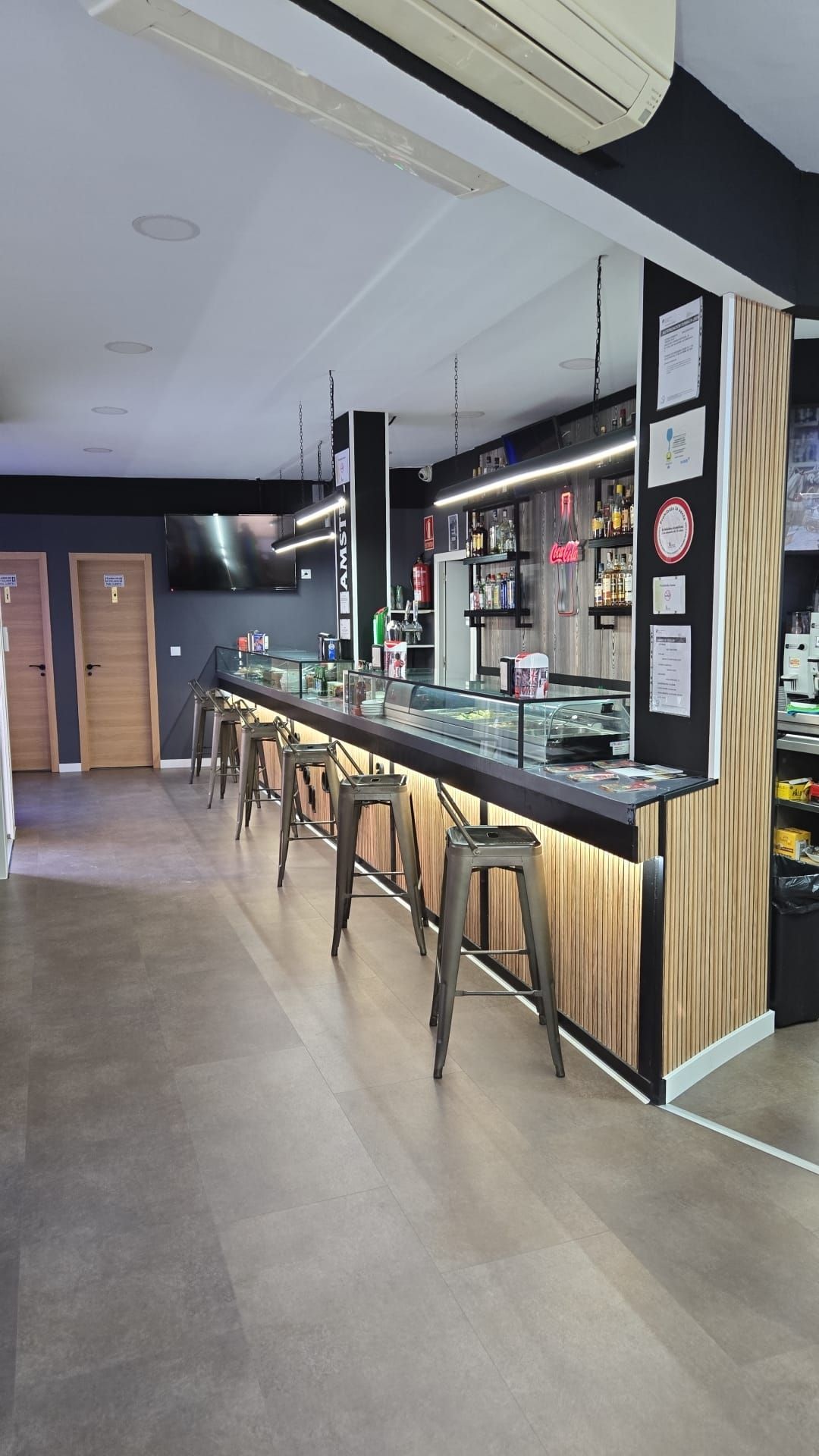 Barra con taburetes, encimera con exposición de comida y fachada revestida de madera y color negro.