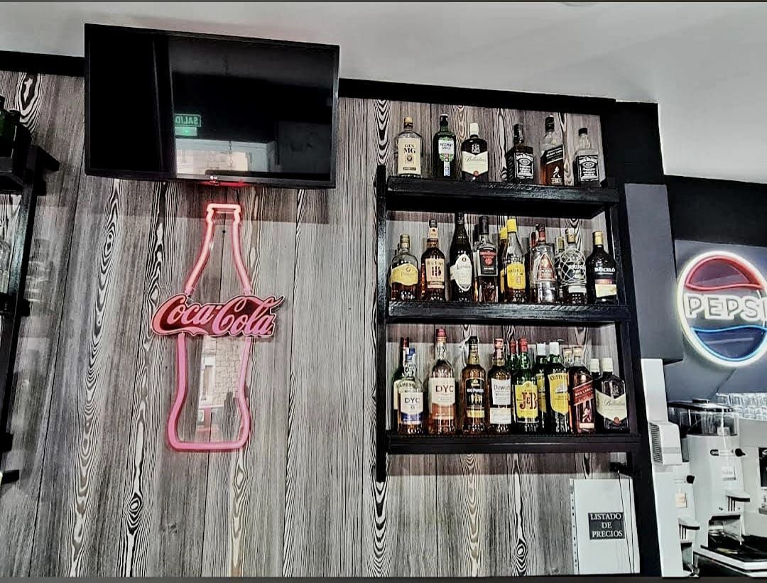 Área de bar con estantes de botellas de licor, un letrero de neón de botella de Coca Cola y un televisor.