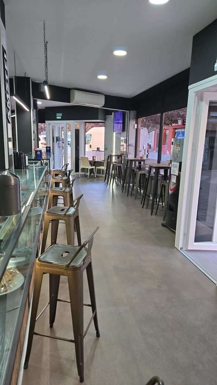 Interior de un bar con taburetes altos en la barra y mesas. Suelo gris, decoración oscura y luz exterior visible.